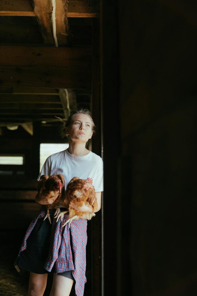 A young woman stands indoors holding two chickens, embracing rural farm life.